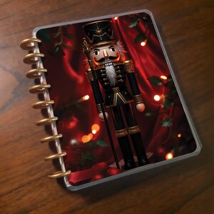 Puede incluir: Una agenda con temática navideña. La portada presenta un detallado soldado cascanueces con uniforme negro y detalles dorados, sosteniendo un bastón. El fondo es rojo intenso con luces suaves y borrosas, sugiriendo un ambiente festivo.