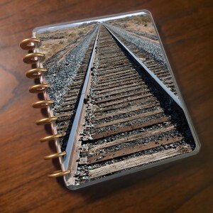 May include: A spiral-bound notebook featuring a photograph of a railroad track receding into the distance. The notebook's binding is gold-colored, and the cover is clear, showcasing the image of the tracks, gravel, and the horizon.