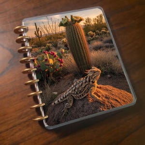 May include: A disc-bound notebook featuring a cover image of a horned lizard resting on a rock in a desert landscape. The scene includes a tall saguaro cactus with yellow blooms and prickly pear cacti with pink fruit under a warm sunset sky.