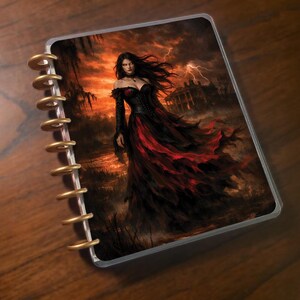 May include: A planner with a fantasy art cover featuring a woman in a black and red gown against a stormy, orange sky. The planner has gold-colored spiral binding and a clear plastic cover.