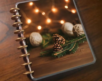 May include: A clear-covered planner with a gold spiral binding. The planner's image features a festive arrangement of pinecones, greenery, and string lights on a wooden surface, creating a warm, seasonal theme.