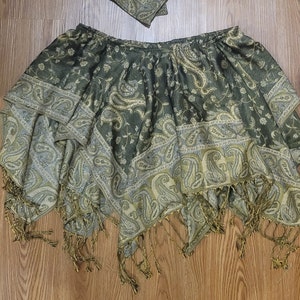May include: An olive green and cream colored skirt featuring a detailed paisley pattern throughout. The garment has an elasticized waistband, a layered handkerchief hemline, and decorative fringe trim along the bottom edges of the fabric.