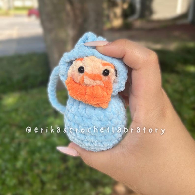 May include: A handmade, light blue crochet gnome with an orange beard and a matching hood. The gnome has black eyes and a loop for hanging. The text "@erikascrochetlabratory" is visible.