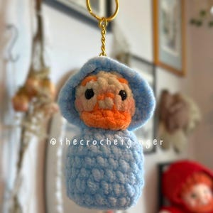 May include: A handmade crochet keychain featuring a duck character with a light blue hooded outfit and an orange beak. The keychain has a gold-colored ring. The text "@thecrochetginger" is visible on the bottom of the keychain.
