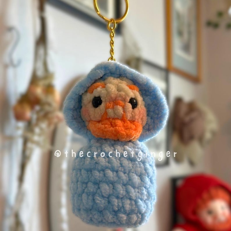 May include: A handmade crochet keychain featuring a duck character with a light blue hooded outfit and an orange beak. The keychain has a gold-colored ring. The text "@thecrochetginger" is visible on the bottom of the keychain.