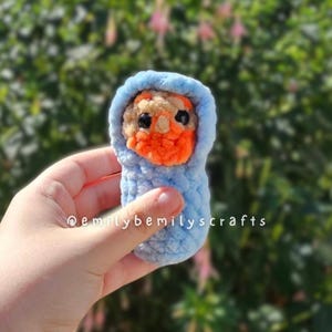 May include: A handmade crochet baby doll swaddled in a light blue blanket. The doll has an orange face and a light brown head, with black eyes. The text "@emilybemilyscrafts" is visible.