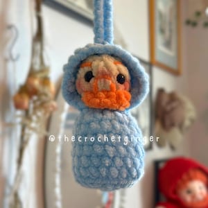 May include: A handmade crochet duckling in a light blue hooded outfit. The duckling has an orange beak and is wearing a blue hood. The text "@thecrochetginger" is visible on the bottom of the image. The duckling is hanging from a blue loop.