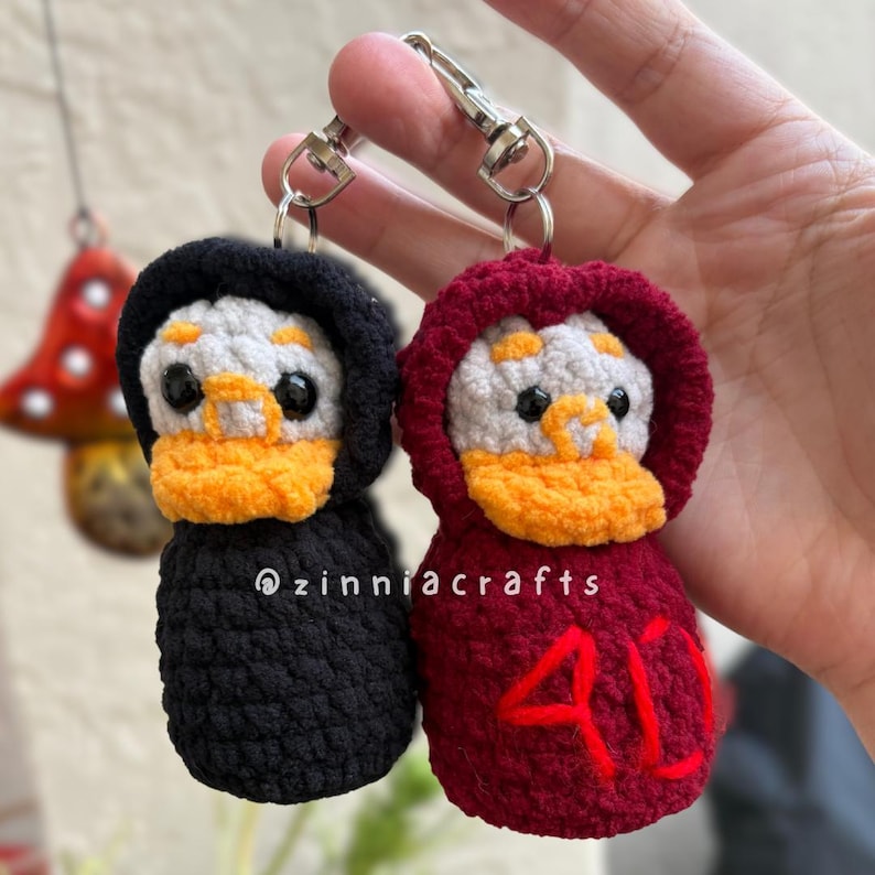 May include: Two handmade crochet duck keychains. One is black, the other is burgundy, both with white and orange faces. The burgundy duck has the number 40 in red. Each keychain has a silver clasp. The text "@zinniacrafts" is visible.