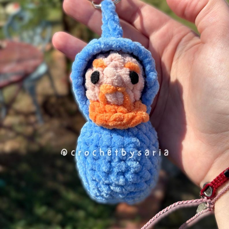 May include: A handmade blue crochet keychain featuring a gnome-like figure with a pink face, orange beard, and black eyes. The keychain has a blue hood and body. The text "@crochetbysaria" is visible on the bottom.