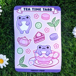 May include: A sticker sheet with a purple border featuring the text "TEA TIME TARO" and "FROGGY POOF". The sheet includes illustrations of a frog, teacup, teapot, macarons, cake, flowers, and leaves.