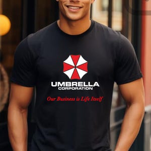 May include: Black crew neck t-shirt featuring the Umbrella Corporation logo in red, white, and black. The text "UMBRELLA CORPORATION" is above the text "Our Business is Life Itself" in red.