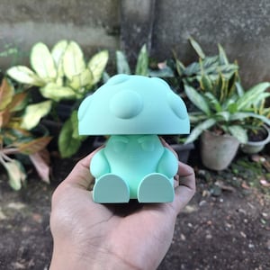 May include: A light green mushroom-shaped figurine with a smiling face, arms, and legs. The mushroom cap has several raised circular details. The figurine is held in a hand, with various potted plants in the background.
