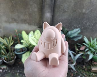 May include: A light peach-coloured figurine of a cartoonish creature with one eye, two ears, and a wide grin. The figure is sitting with its legs extended. The background is blurred with green plants.