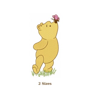 Op de afbeelding: Geborduurd beeld van een gele beer met een roze vlinder op zijn neus, staand op groen gras. De tekst "2 Sizes" staat onder de afbeelding.