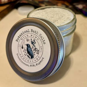 May include: A silver jar with a white label that reads "Botanical Bath Salts Wild Wing Apothecary Frankincense, MSM, Mugwort". The jar is filled with white bath salts.