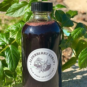 May include: A glass bottle of dark purple elderberry syrup with a white label that reads "Elderberry Syrup - Wild Wing Apothecary".