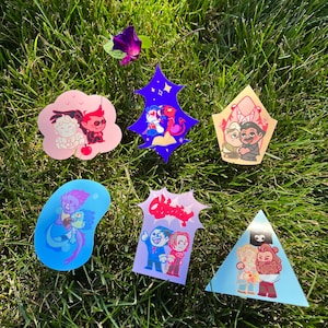 May include: Six colorful stickers featuring cartoon couples. The stickers are on a green grassy surface. The stickers feature a variety of characters and designs, including a couple holding hands, a couple with a dragon, a couple with a crown, a couple with a heart, a couple with a fish, and a couple with a speech bubble.