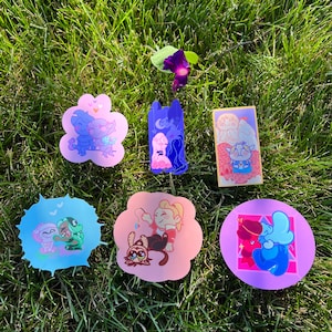 May include: Six colorful stickers featuring cartoon couples. The stickers are on a green grassy surface. The stickers are shaped like a flower, a rectangle, a circle, and a cloud.