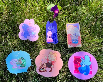 May include: Six colorful stickers featuring cartoon couples. The stickers are on a green grassy surface. The stickers are shaped like a flower, a rectangle, a circle, and a cloud.