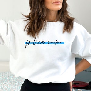 May include: White sweatshirt with the text "police mom" in black script with a thin blue line above the text.