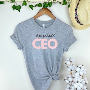 May include: A heather gray t-shirt with rolled-up sleeves. The shirt features the words "household CEO" in a black script font above the word "CEO" in a large, light pink block font. A straw hat with a floral band hangs in the background.