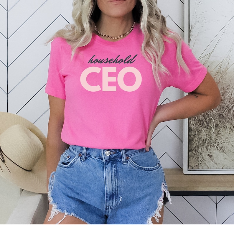 May include: A bright pink t-shirt with the words "household CEO" printed on the front in a contrasting light pink color. The text is in a playful font. The shirt is a crew neck style and appears to be made of a soft material.
