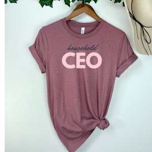 May include: A mauve-colored t-shirt with rolled-up sleeves. The shirt features the words "household CEO" in a two-tone design. The word "household" is in a dark gray font, and the word "CEO" is in a light pink font. The shirt is hanging on a wooden hanger.