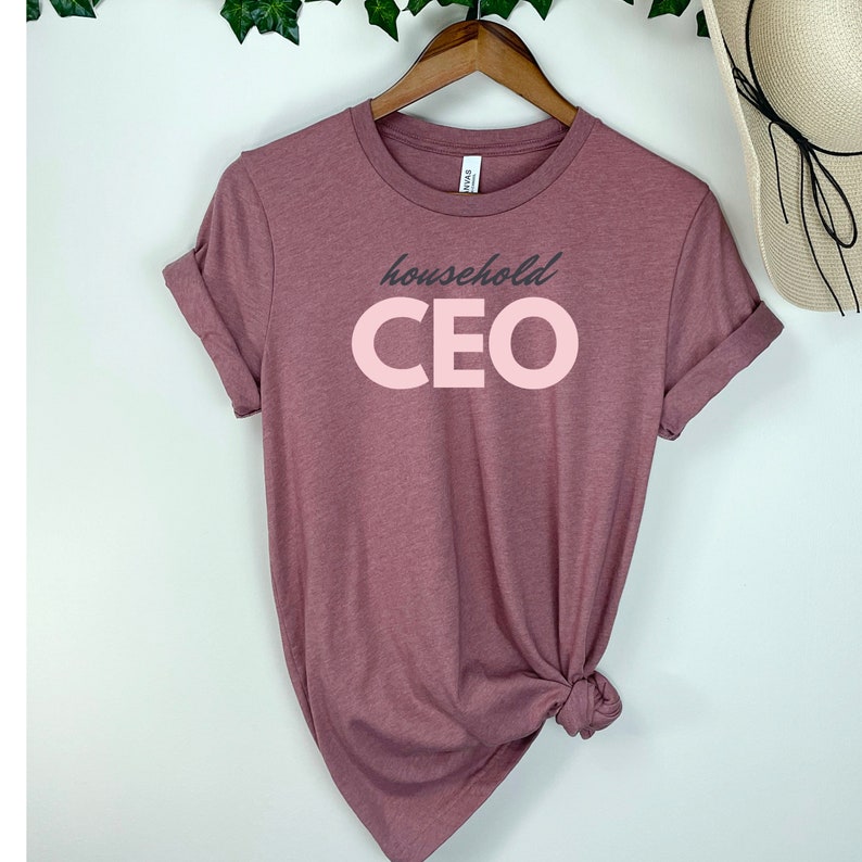 May include: A mauve-colored t-shirt with rolled-up sleeves. The shirt features the words "household CEO" in a two-tone design. The word "household" is in a dark gray font, and the word "CEO" is in a light pink font. The shirt is hanging on a wooden hanger.
