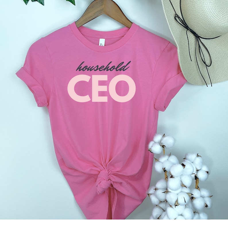 May include: A bright pink t-shirt with the words "household CEO" printed on the front in a contrasting light pink and dark gray font. The t-shirt is knotted at the waist and hangs on a wooden hanger.