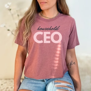 May include: A mauve-colored t-shirt with the words "household CEO" printed on the front in a two-tone design. The word "household" is in a dark gray script, and "CEO" is in a light pink, bold, sans-serif font. The shirt has short sleeves.