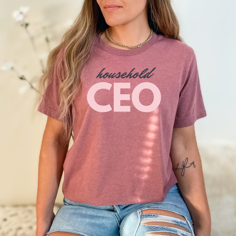 May include: A mauve-colored t-shirt with the words "household CEO" printed on the front in a two-tone design. The word "household" is in a dark gray script, and "CEO" is in a light pink, bold, sans-serif font. The shirt has short sleeves.