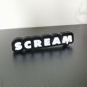 Könnte beinhalten: Ein schwarz-weißes Schild mit der Aufschrift "SCREAM". Das Wort ist weiß, schwarz umrandet und hat eine abgerundete, blasenartige Schriftart. Das Schild befindet sich auf einer dunkelgrauen Oberfläche.