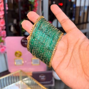 May include: A stack of emerald green bangles featuring a translucent finish with embedded gold glitter particles. The set is framed by two textured gold-toned metal bangles on the outer edges, creating a cohesive and layered circular wrist accessory.