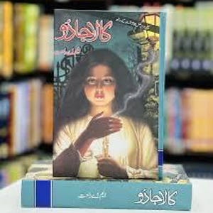 May include: A hardcover book titled "The Wizard" in Arabic, featuring a woman holding a glowing object. The cover art is a painting with a teal and blue background. The book is on top of another book.