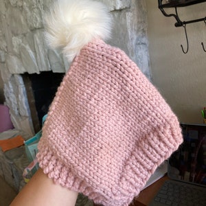 May include: A pink knitted hat with a white faux fur pom pom on top. The hat has a long tie that hangs down the side.