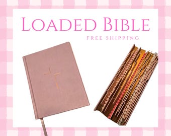 Puede incluir: Una "LOADED BIBLE" rosa con una cruz dorada en la portada y una sección del libro abierto que muestra cuentas de colores. El libro tiene una cinta rosa como marcador. El fondo es un patrón de cuadros rosas y blancos.