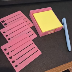 May include: A set of three pink plastic stencils with different cutout shapes including squares, circles, and hearts, alongside a pink plastic holder containing a stack of yellow square sticky notes and a light blue retractable ballpoint pen.