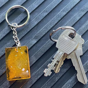 May include: A silver keyring with a rectangular, amber-colored resin keychain. The keychain has embedded glitter and is attached to a chain. Next to the keychain are three silver keys with the word "Master" on them.