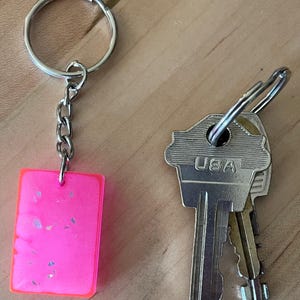 May include: A bright pink rectangular keychain with silver glitter accents, attached to a silver chain and ring. Two keys with the text "USA" are next to the keychain on a wooden surface.