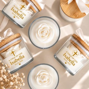 Puede incluir: Velas de cristal con tapas de bambú y cintas blancas, con un icono de cruz dorada, el texto A CHILD OF GOD, el nombre Isabella y la fecha 03.23.2025. Dos velas están abiertas, revelando cera blanca con forma de rosa en el centro.