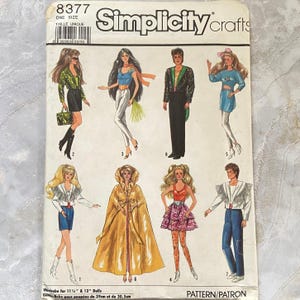 May include: A Simplicity Crafts sewing pattern envelope numbered 8377 for 11.5 and 12 inch dolls. The front features illustrations of seven different doll outfits, including jackets, skirts, pants, and a long gold cape, with text labels and a barcode.