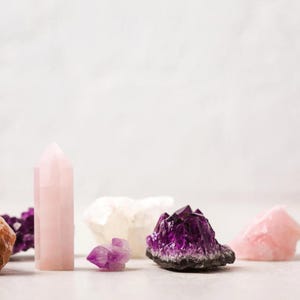Op de afbeelding: Een verzameling roze en paarse kristallen, waaronder een rozenkwarts toren, een amethistcluster en een kleinere amethistpunt. De kristallen zijn gerangschikt op een wit oppervlak.