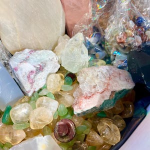 Op de afbeelding: Een verzameling van verschillende kristallen en stenen, waaronder helder kwarts, rozenkwarts, amethist en groene aventurijn. De kristallen zijn gerangschikt in een decoratieve kom, met enkele stenen die uit de kom vallen. De kristallen zijn in verschillende vormen en maten aanwezig, en sommige hebben een natuurlijke, ruwe textuur.