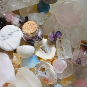 Op de afbeelding: Een kom vol met verschillende kristallen en stenen, waaronder helder kwarts, rozenkwarts, amethist en witte agaat. Twee kleine glazen potjes met kurken deksels bevatten kleinere kristallen.
