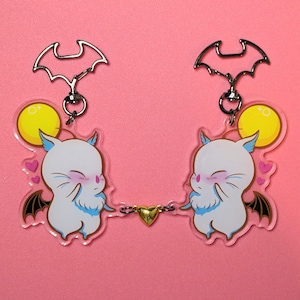 May include: Two acrylic keychains linked by a gold heart charm. Each keychain depicts a white cartoon bat-like creature with closed eyes, a yellow moon, and pink hearts. The keychains have silver bat-shaped attachments.