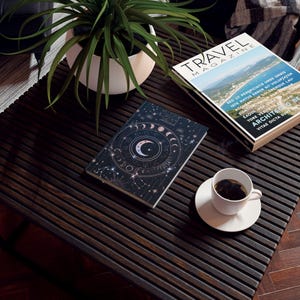 Op de afbeelding: Een donkerblauw notitieboek met een gouden en roze hemelse ontwerp. Het notitieboek ligt open op een donkerhouten tafel met een kop koffie en een reismagazine.