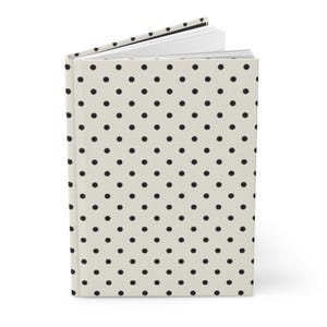 May include: A hardback journal with a cream-coloured cover featuring a repeating pattern of small black polka dots. The book is shown slightly open, revealing white pages inside. The binding is visible along the left side of the upright book.
