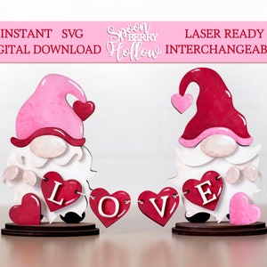 Könnte beinhalten: Zwei Gnome halten ein Banner mit der Aufschrift "LOVE" mit rosa und roten Herzen. Die Gnome tragen rosa und rote Hüte mit Herzen darauf. Die Gnome stehen auf Holzsockeln.