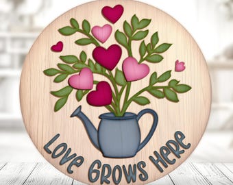 May include: A wooden round sign with a blue watering can filled with pink and red hearts on green stems. The sign says "Love Grows Here" in white lettering.