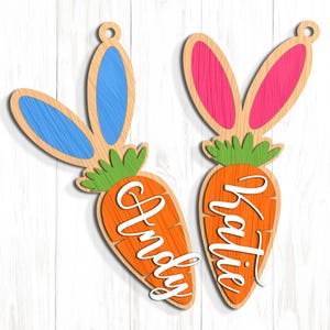 May include: Two wooden carrot-shaped ornaments with bunny ears. The carrots are orange with green tops and have the names "Katie" and "Andy" written on them. The bunny ears are pink and blue.
